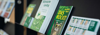 Im Vordergrund liegen verschiedene Broschüren auf einem Regal, im Hintergrund erkennt man weitere Regale mit Publikationen darauf.