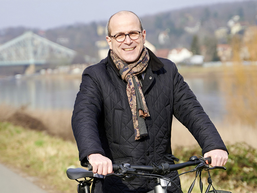 Porträt des Abgeordneten Martin Modschiedler an der Elbe mit den Händen am Fahrradlenker. Im Hintergrund ist das Blaue Wunder erkennbar.