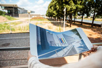 Jemand hält den Plan mit dem Erweiterungsbau in der Hand. Im Hintegrund ist die Freifläche zu sehen, auf der der Bau entstehen wird.