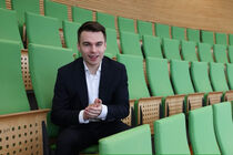 Ein junger Mann in schwarzem Anzug sitzt auf einer Besuchertribüne mit grünen Klappstühlen.