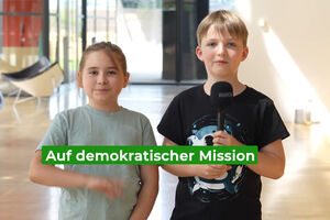 Zwei Kinder stehen mit den Mikrofon in der Lobby und schauen lächelnd in die Kamera.