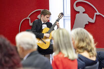 Ein junger Mann spielt Gitarre. Einige Personen sind von hinten zu sehen, während sie ihm zuhören.